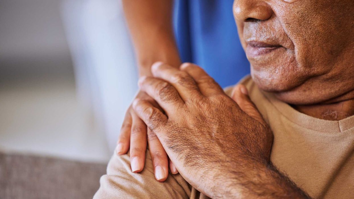 A close up of a nurse's hand resting on the shoulder of an older man who's hand rests on top.