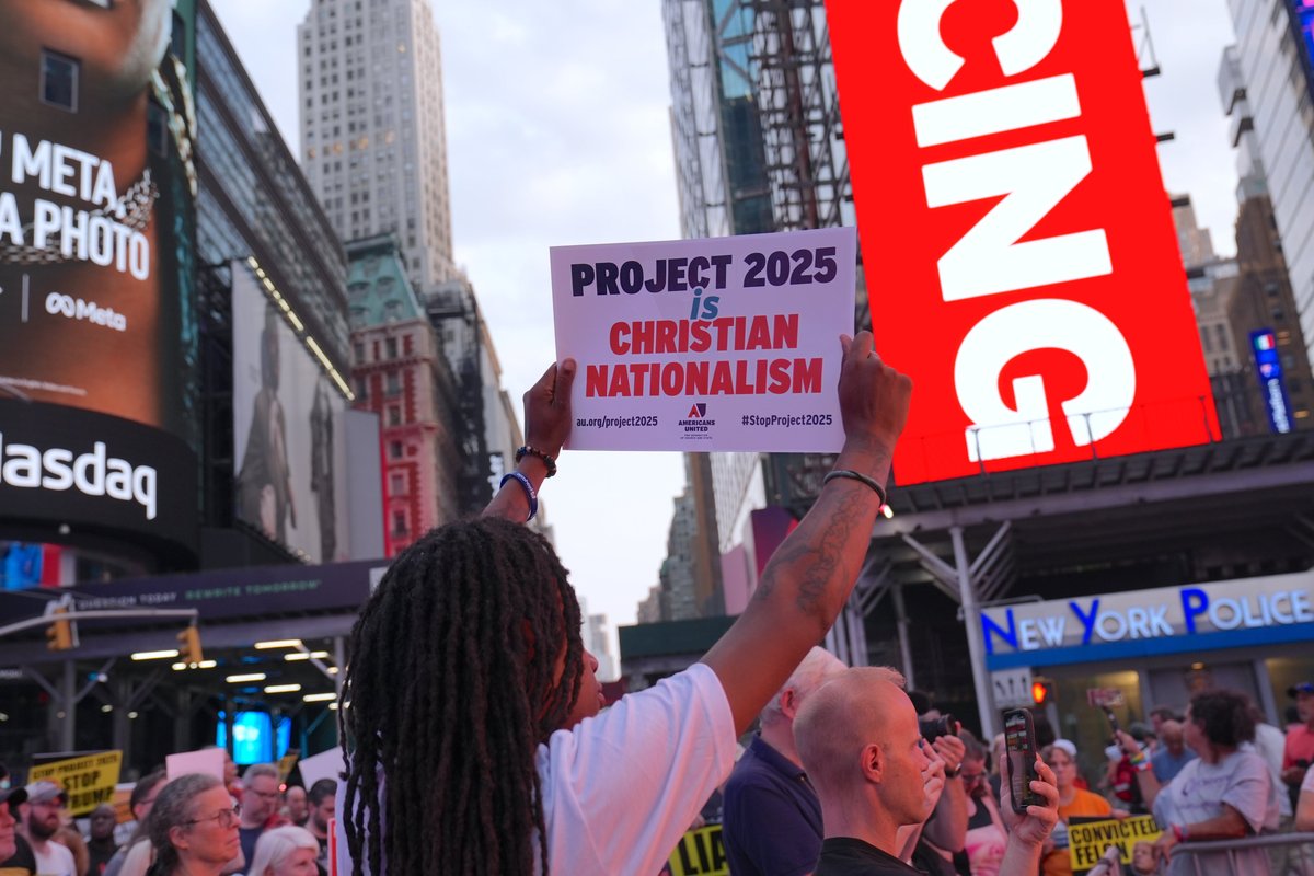 A crowd of protesters in Times Square, New York City, with one person holding a sign that reads "PROJECT 2025 is CHRISTIAN NATIONALISM" by Americans United for Separation of Church and State. The sign includes the hashtags #StopProject2025 and au.org/project2025. The background features prominent advertisements, including a Meta billboard and the Nasdaq building.