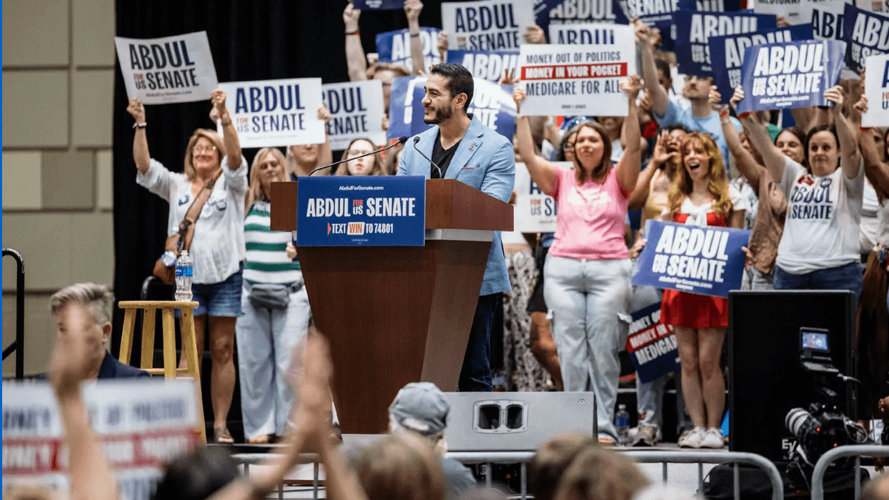 A crowd of supporters holding signs behind Senate candidate Dr. Abdul El-Sayed as he stands and speaks at a podium outside in front of a crowd.