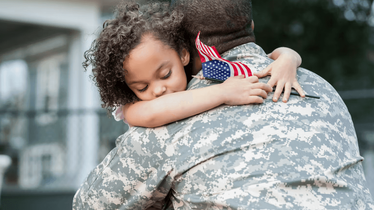 A person in a military uniform hugging a child, who is hugging them back with a small U.S. flag in her hand.
