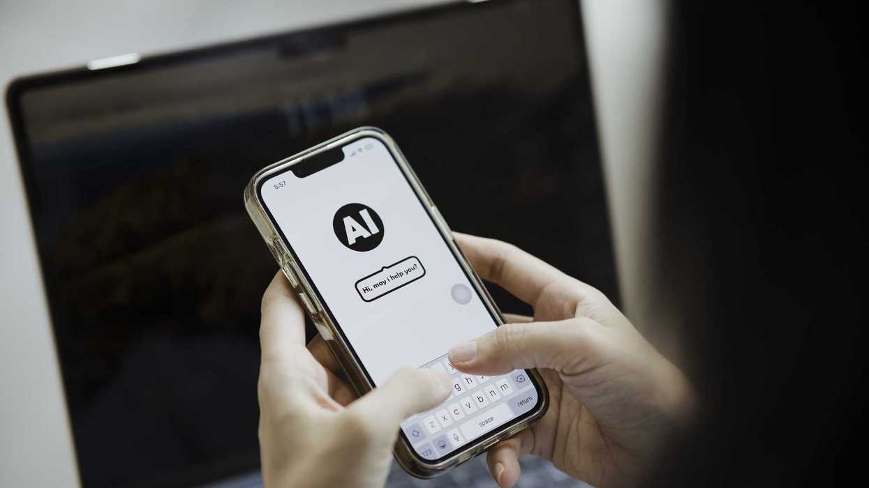 A person interacts with an AI chatbot on a smartphone. The screen displays a conversation interface with the text "Hi, may I help you?" suggesting the use of an AI assistant app. A laptop is visible in the background, emphasizing a tech-savvy environment.