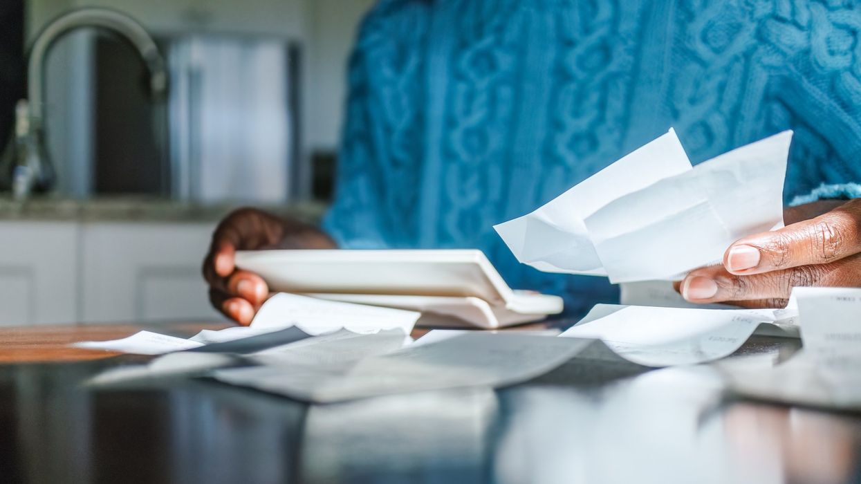 A person sits in their kitchen, holding and looking at receipts in one hand, with a calculator in their other hand.