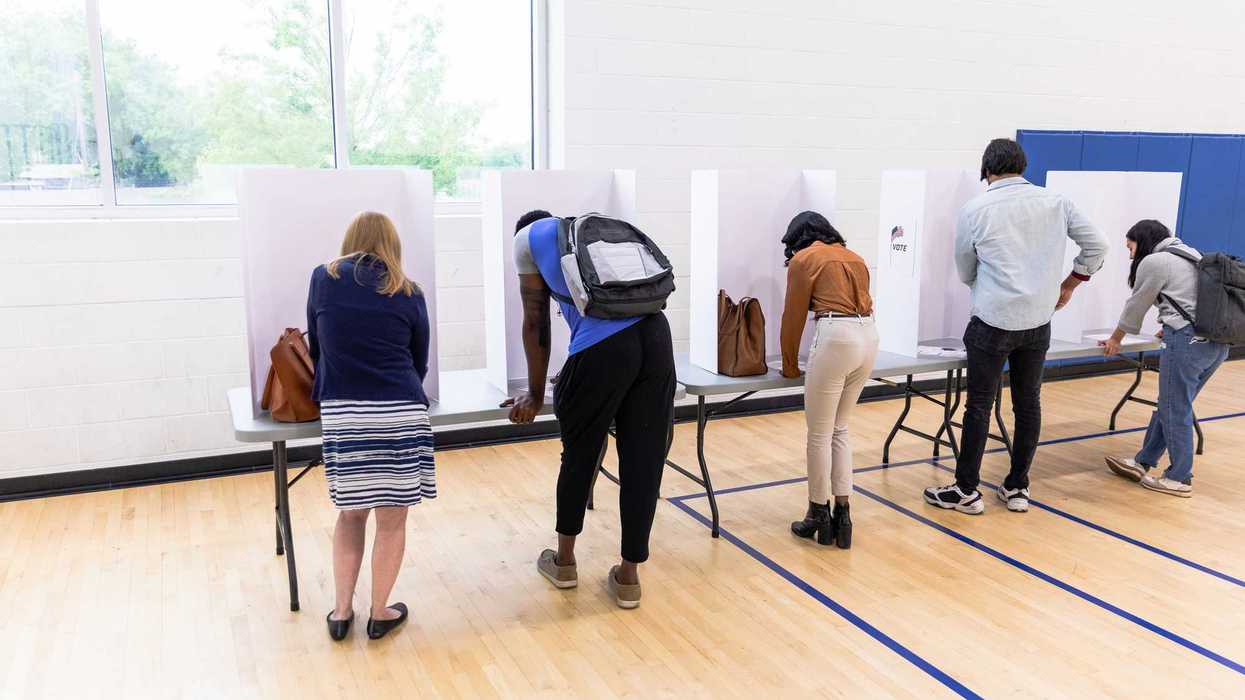 A rear view photo of a multiracial group of people writing their choices on their ballots in private voting booths set up on a long table.