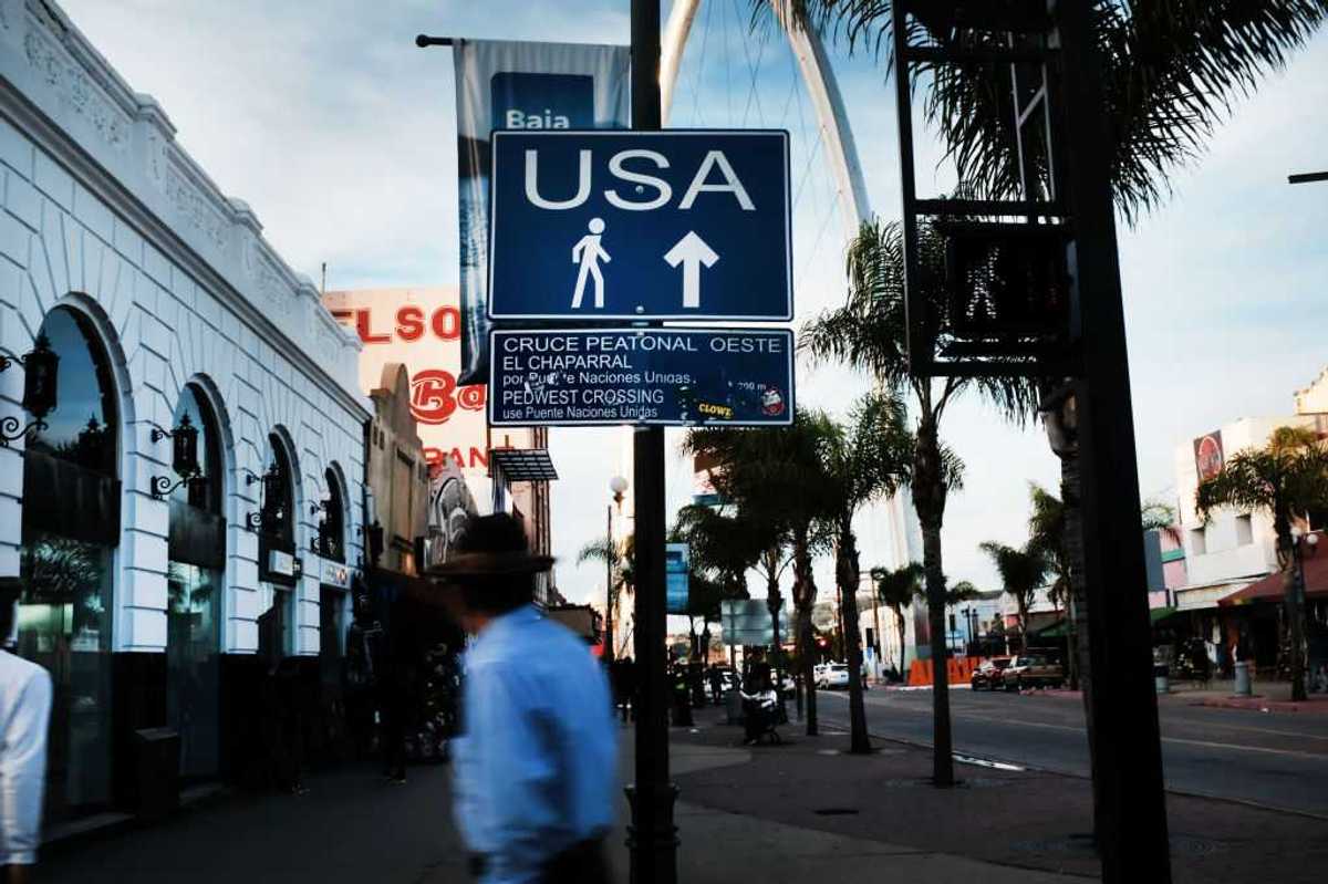 A sign directs pedestrians towards the United States in Tijuana, Mexico. A person walks in the foreground.