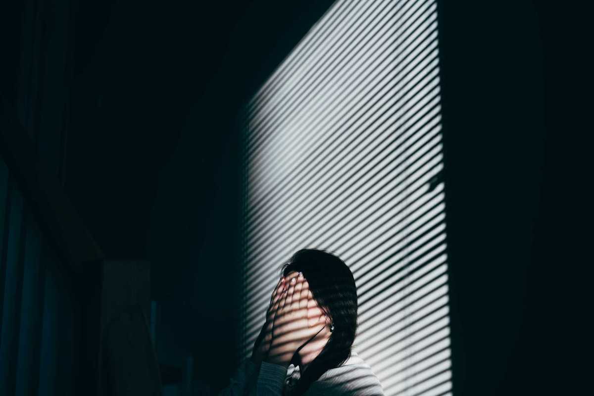 A solemn portrait of a woman looking down, bathed in dramatic light and shadow from window blinds, emphasizing solitude and deep thought in monochrome.