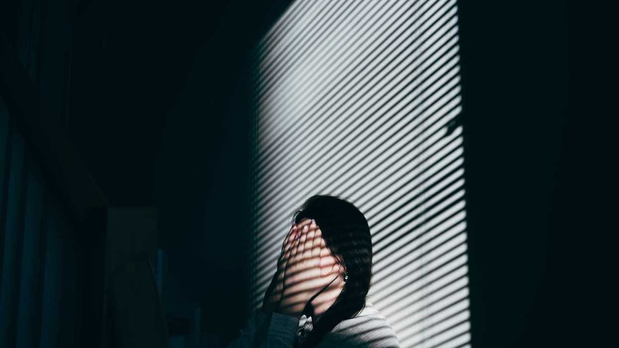 A solemn portrait of a woman looking down, bathed in dramatic light and shadow from window blinds, emphasizing solitude and deep thought in monochrome.