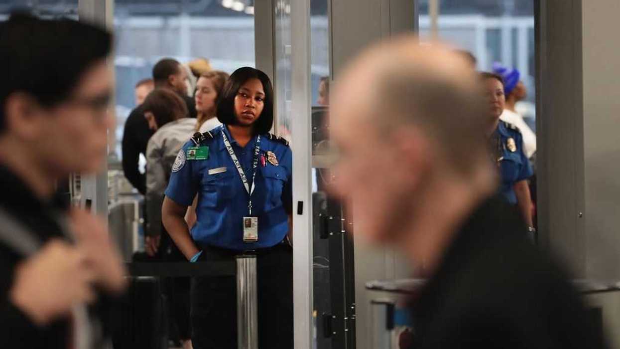 A TSA employee standing in the airport, with two travelers in the foreground.