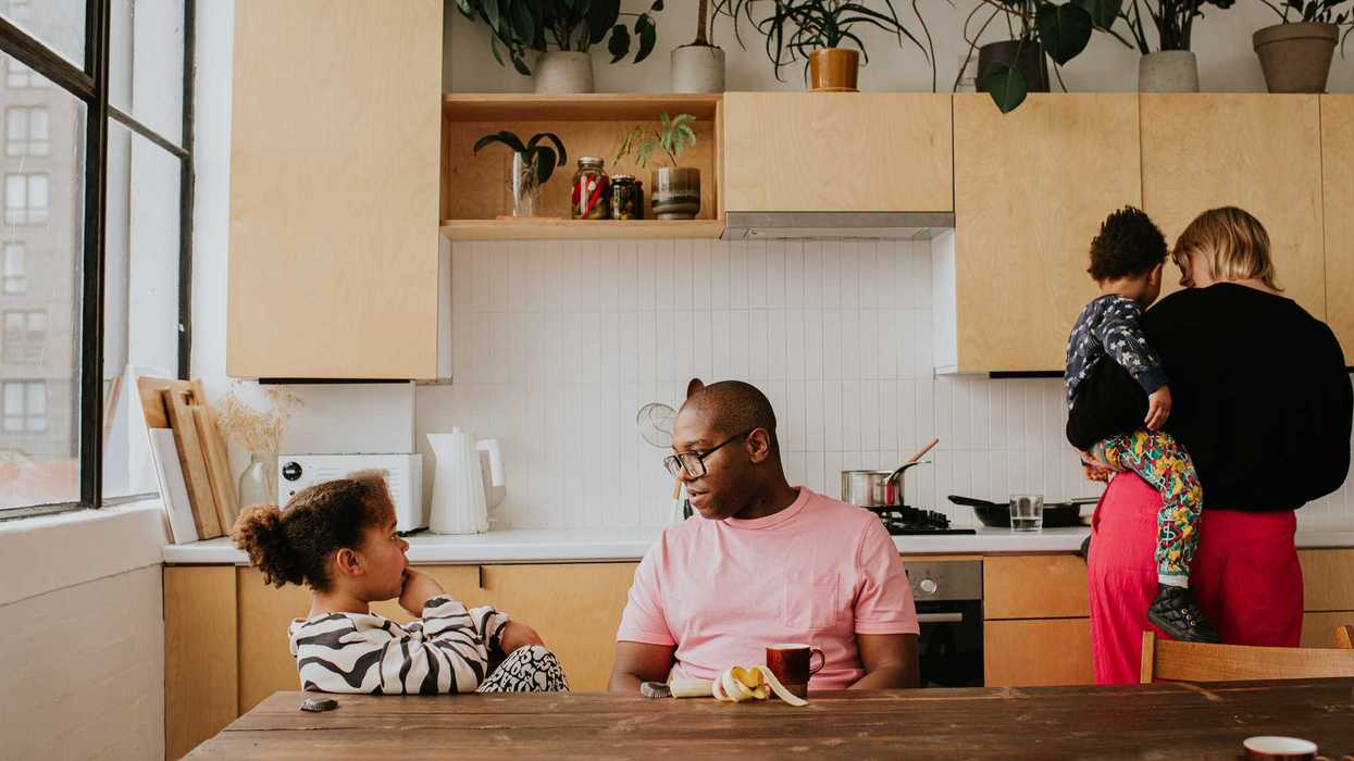 A young family sit at a long dining table in a stylish domestic kitchen. A father and daughter have a discussion at the table, whilst a mother holds her toddler on her hip and prepares a meal on a marble surface. Image depicts busy family life.