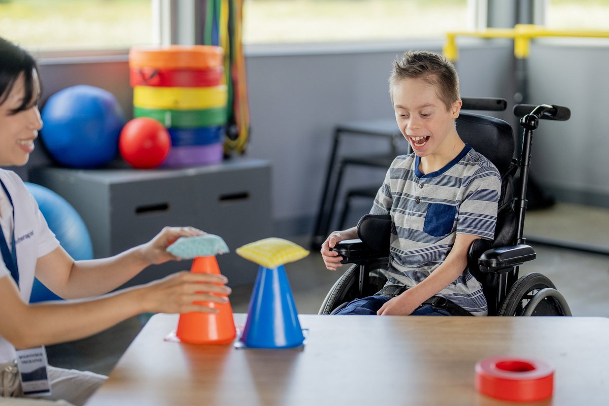 An occupational therapist sits with a young boy at a table as they work on some of his motor skills.