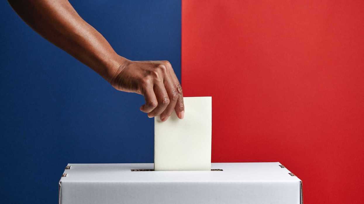 Cardboard ballot box with a voter slip being placed into it by a hand, with a blue and red coloured background.