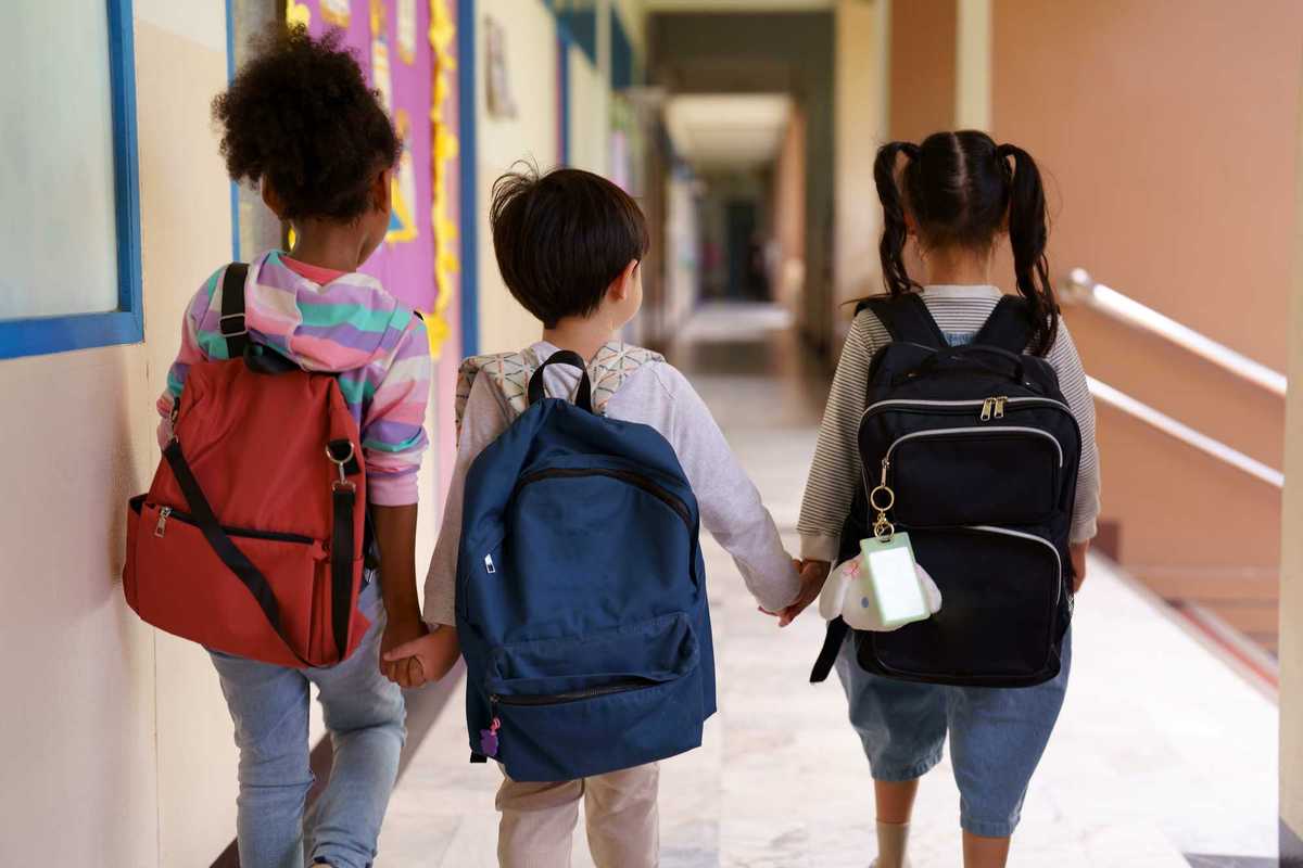 Children holding hands, walking down a school hallway with backpacks on.