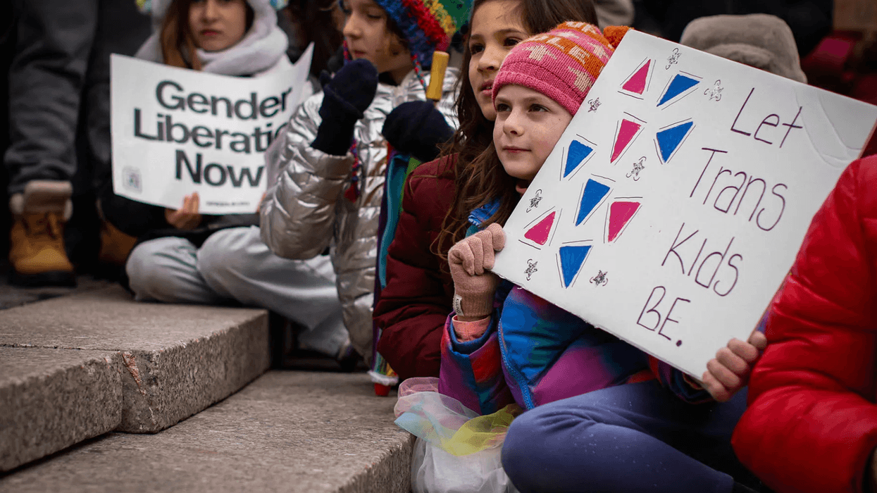 Children sitting down, holding signs that read, "Let Trans Kids Be," and "Gender Liberation Now."