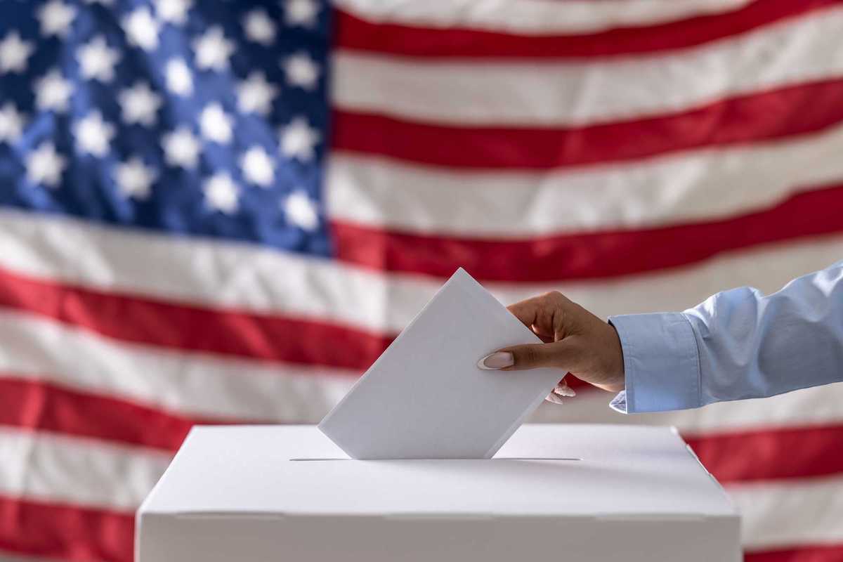 Hand placing a ballot into a box with American flag in the background