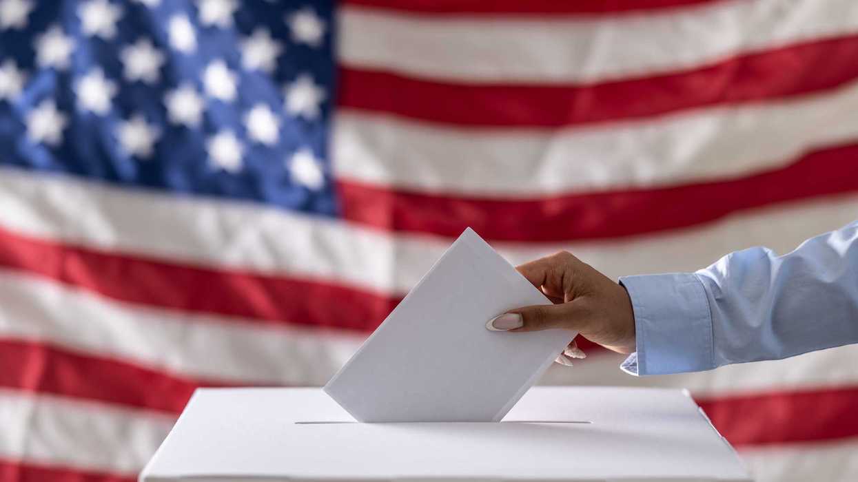Hand placing a ballot into a box with American flag in the background