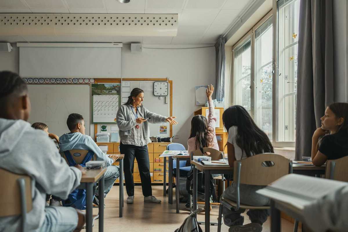 Happy teacher asking girl with hand raised sitting in classroom at elementary school