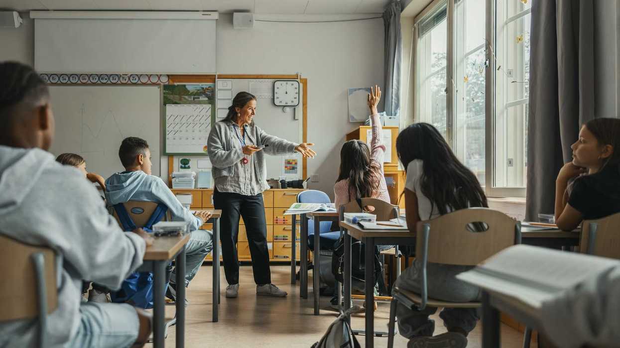 Happy teacher asking girl with hand raised sitting in classroom at elementary school
