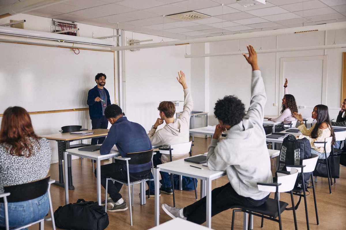 Highschoolers in a classroom, sitting down at their desks and raising their hands.