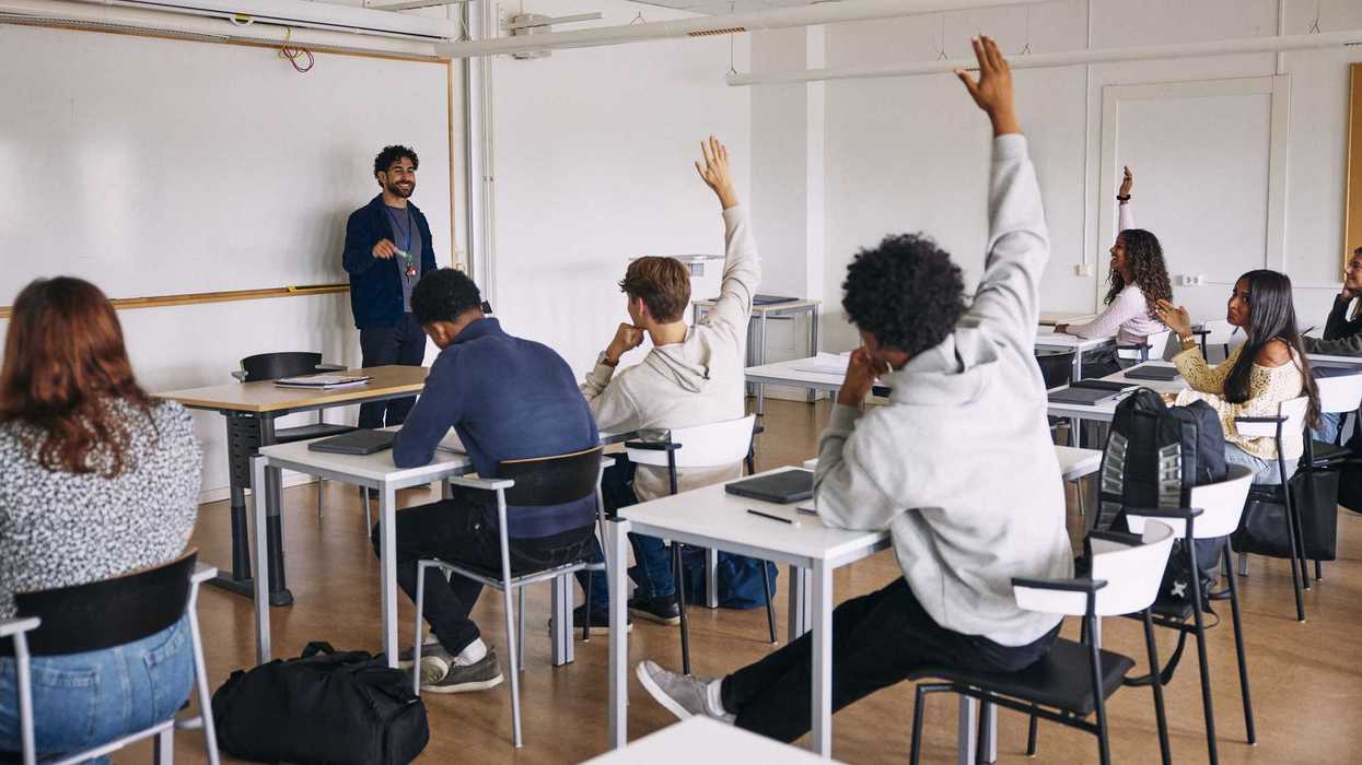 Highschoolers in a classroom, sitting down at their desks and raising their hands.