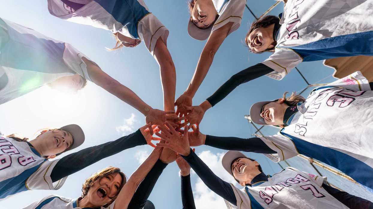 Members of a women's softball team cheerfully join hands during practice on a sunny day, showcasing team spirit, cooperation, and enthusiasm for the sport.