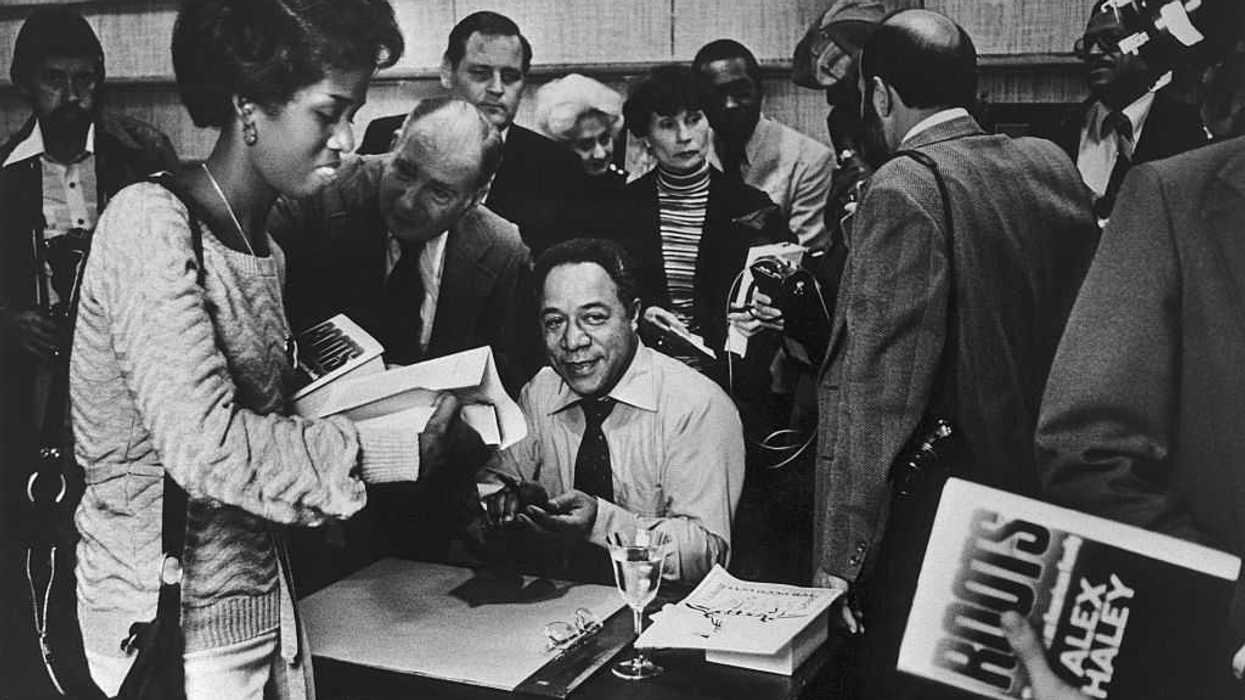 People standing around author Alex Haley, who sits at a desk signing copies of his book, Roots.