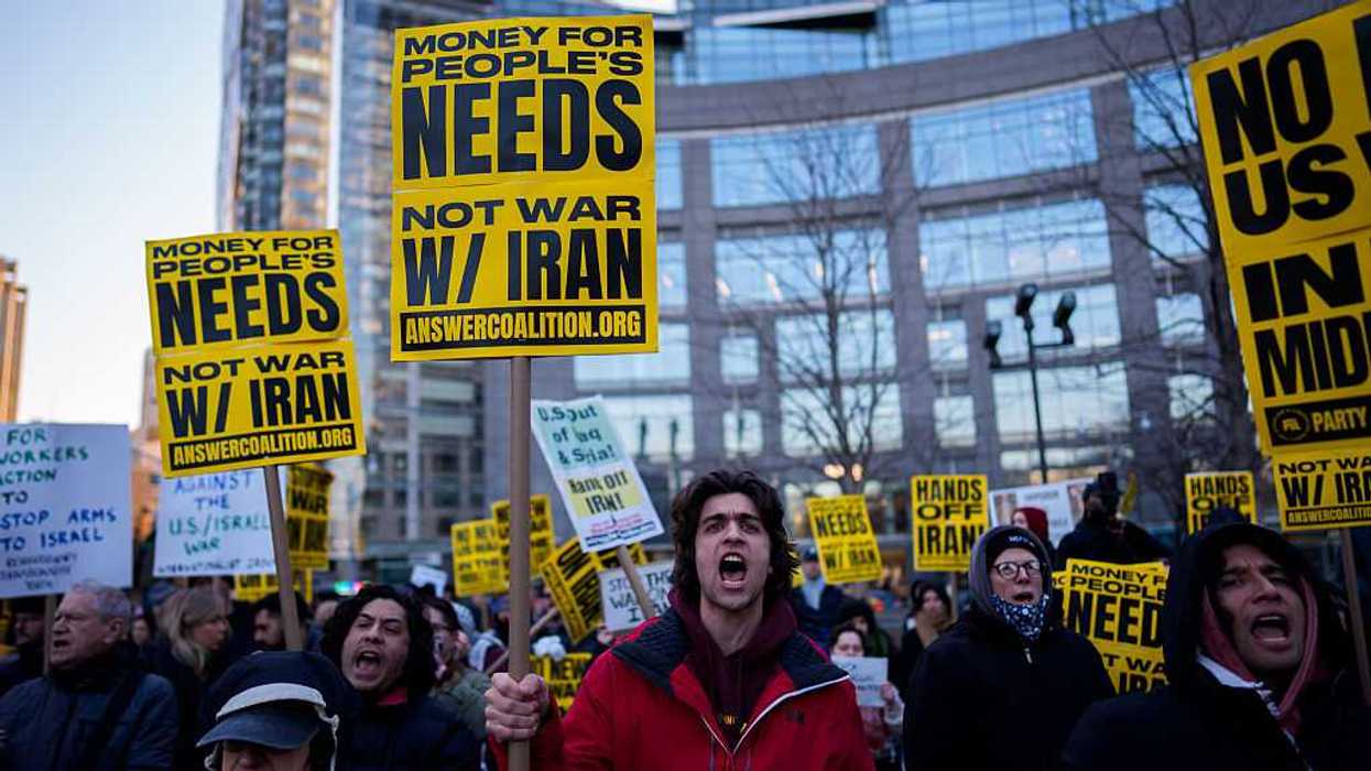 Protestors holding signs that read, "Money for People's Needs, Not War W/ Iran," outside of a building.