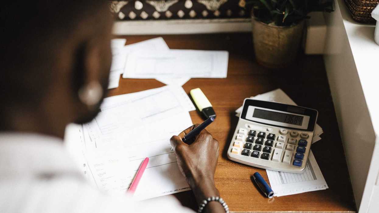 Someone sitting at a desk, writing with a pen on paper, with a calculator and papers by their side.