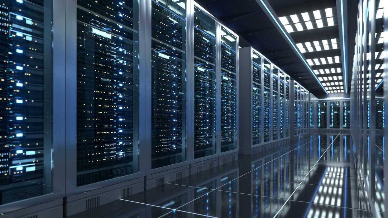 This wide angle shot shows a fully operational modern data center hallway, lined with rows of fully operational server racks behind glass panels. The highly polished reflective floor mirrors the glowing indicator lights and overhead ceiling lights, creating a sleek, futuristic industrial tech environment focused on enterprise cloud computing and digital data storage.