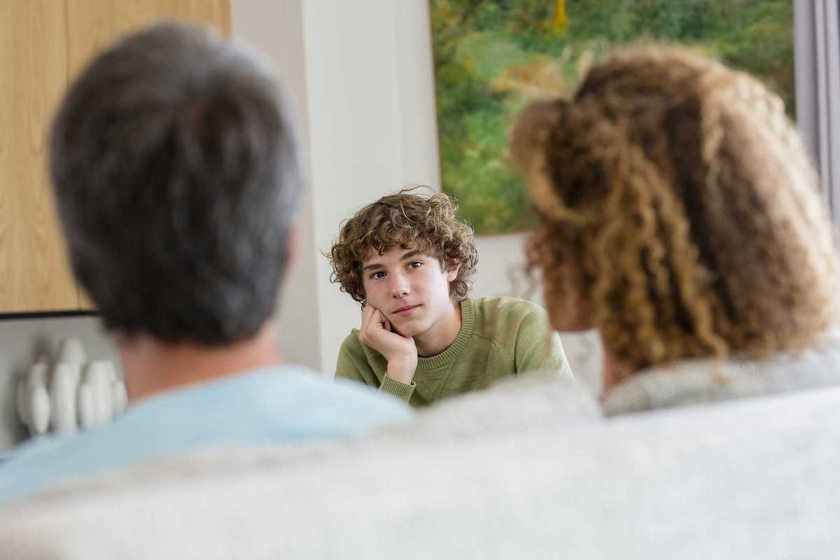 Two parents sitting down on a couch across from their teen son, looking at each other.