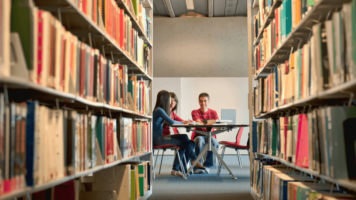 Young adults sitting at a table in a library at the end of an aisle of books.