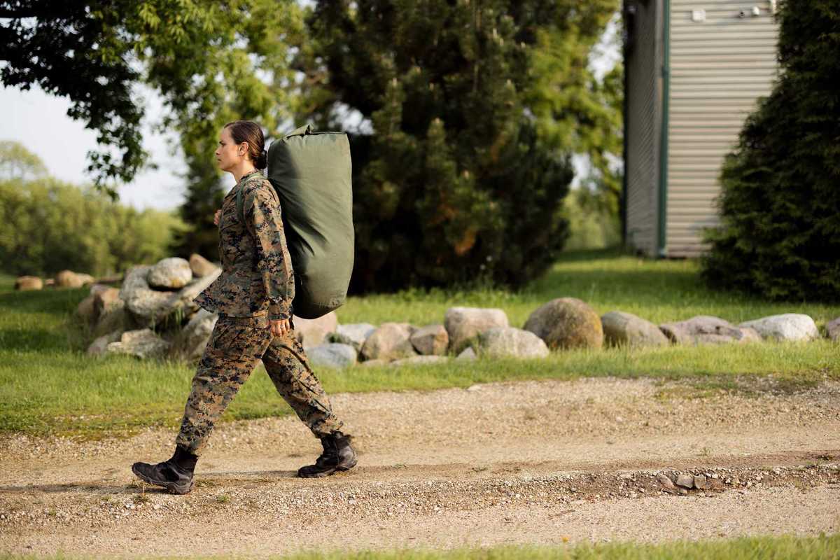 Young female soldier in camouflage uniform strides along a gravel road carrying a heavy duffel bag, focused and determined on mission
