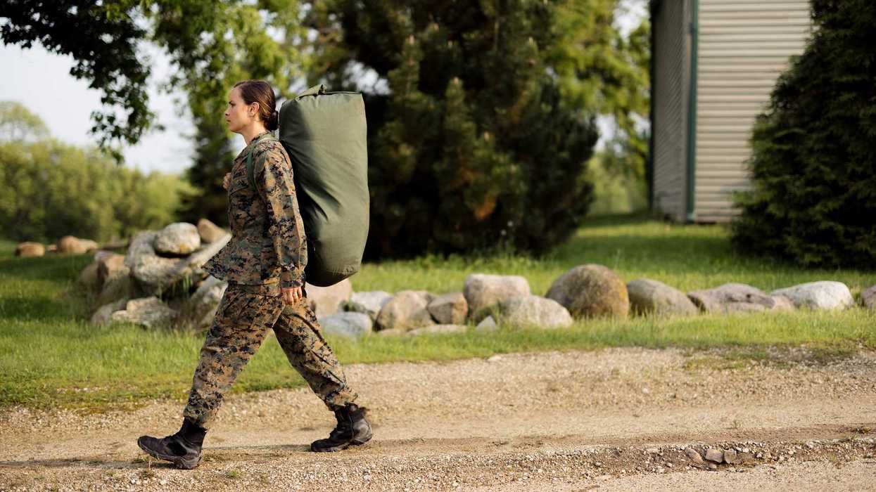 Young female soldier in camouflage uniform strides along a gravel road carrying a heavy duffel bag, focused and determined on mission