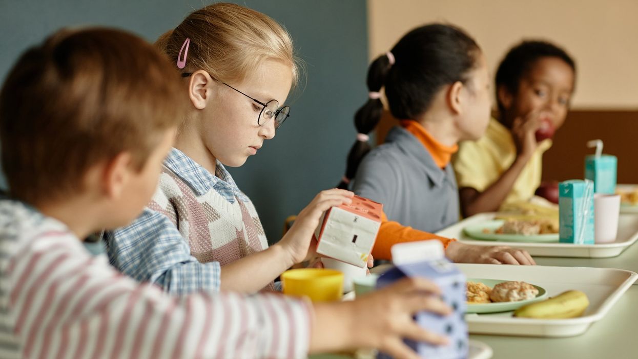 young girl pouring fresh juice or milk into cup sitting at table with classmates while eating lunch in school cafeteria.